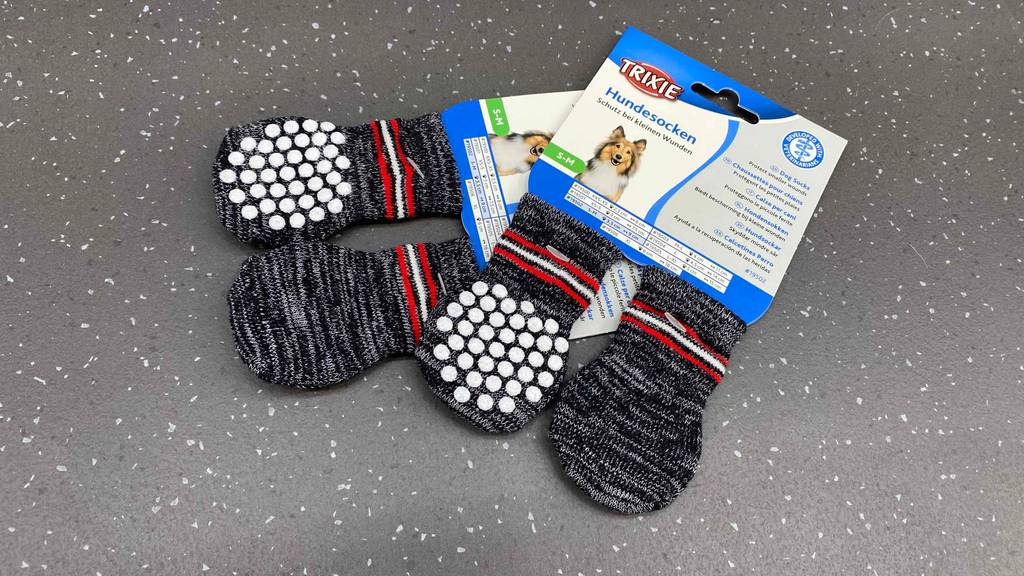 Zwei Paar graue getestete Trixie-Hundesocken liegen original verpackt auf dem Boden.
