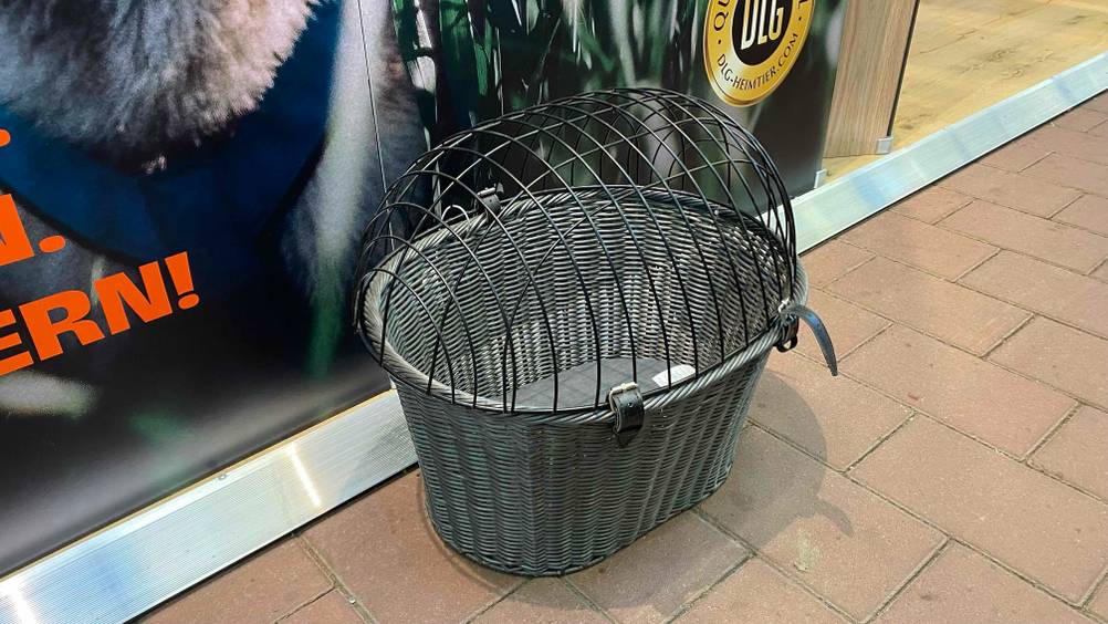 Ein grauer Hundefahrradkorb steht vor einem Plakat auf einem gepflasterten Steinboden, von schräg oben betrachtet.