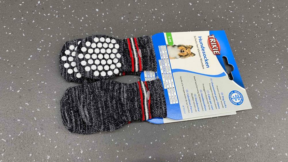 Zwei Paar grau-melierte Trixie-Hundesocken im Test inklusive Gumminoppen befinden sich in ihrer Originalverpackung auf einem grauen Untergrund.