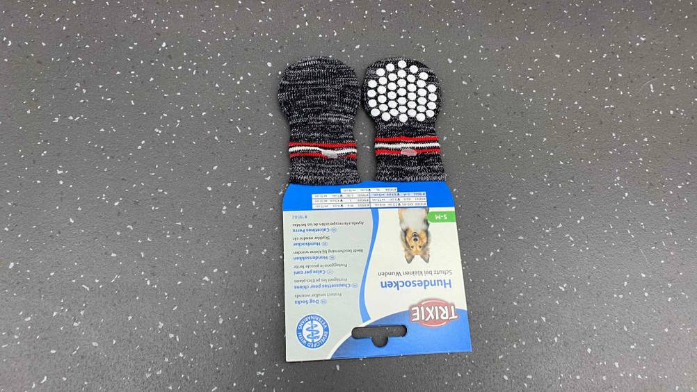 Zwei Paar graue getestete Trixie-Hundesocken liegen in ihrer Originalverpackung auf einem grauen Boden.