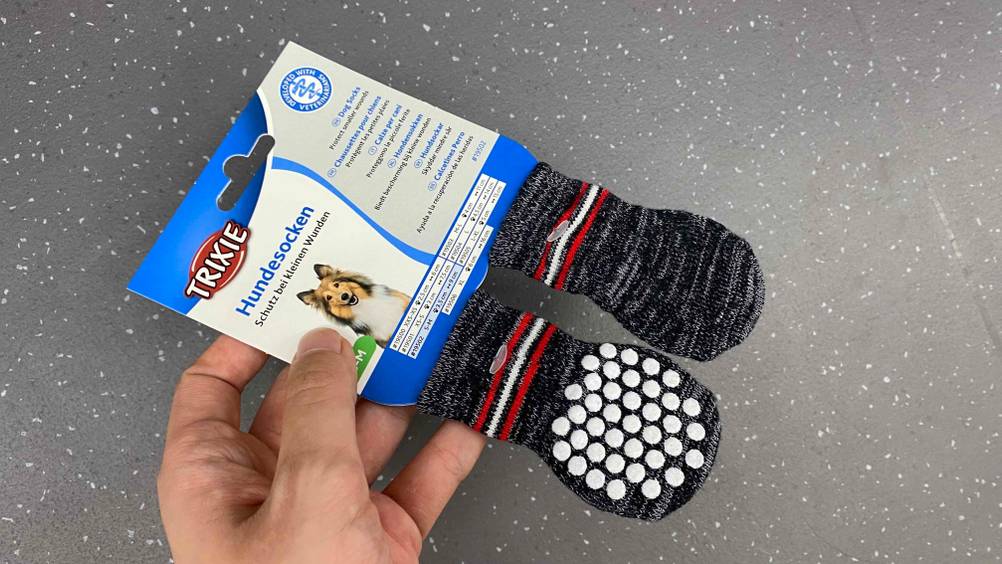 Hand hält graue getestete Trixie-Hundesocken in ihrer Originalverpackung vor grauen Boden.