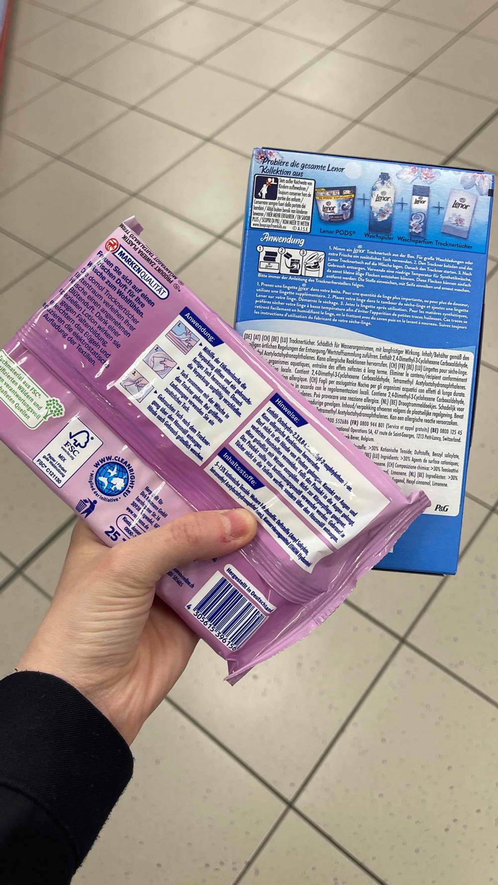 Trockentücher im Test: Eine Hand hält eine violette Kunststofftüte sowie eine blaue Schachtel über einem weißen Fliesenbelag.