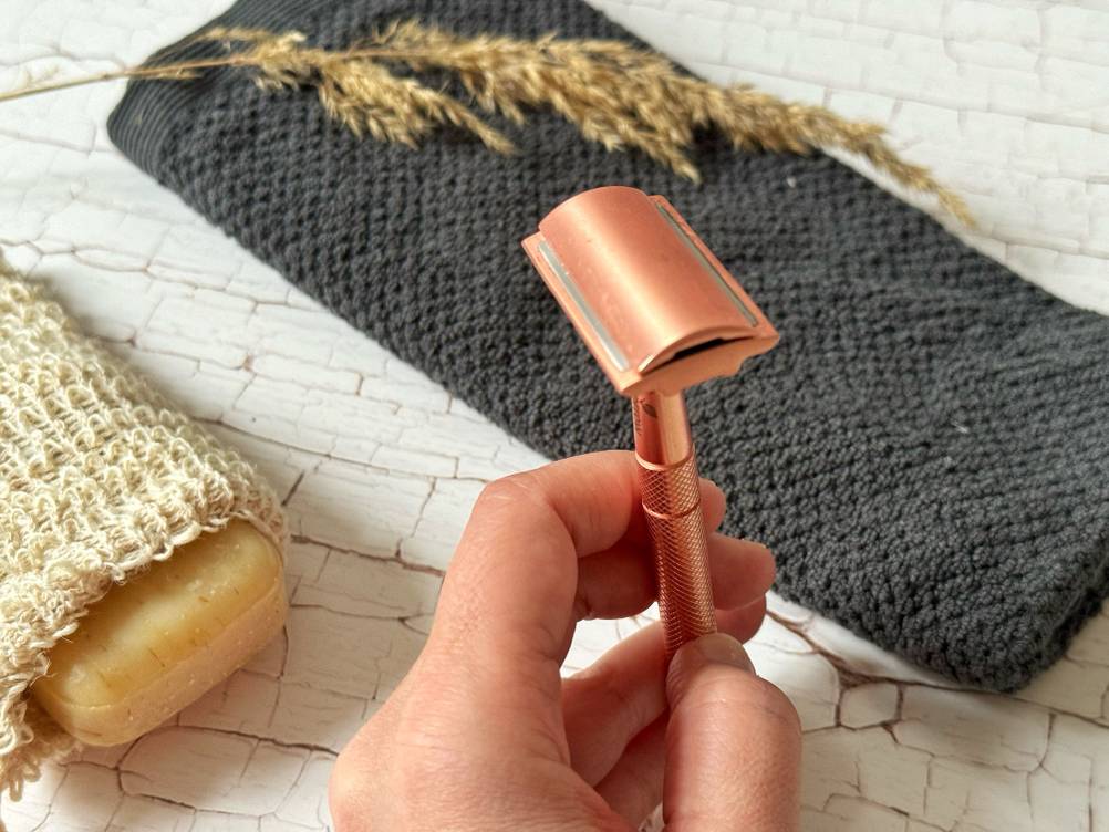 Hand hält einen rosé goldenen getesteten Rasierhobel für Frauen aus Metall, mit Sicht auf die Klinge, über einem grauen Handtuch und einem Stück Natur-Seife.
