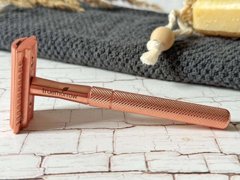 Truemorrow rosé goldener Metall-Rasierhobel für Frauen im Test liegt auf einer weißen Fläche vor einem grauen Handtuch und einem Stück gelber Seife.