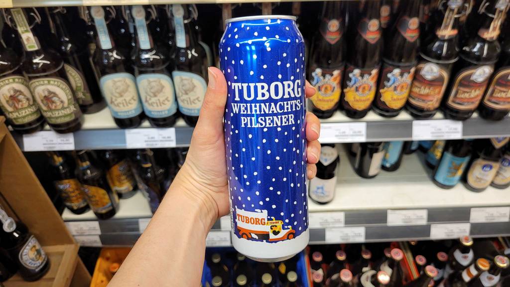 Eine Hand hält eine Dose mit getestetem Weihnachtsbier im Weihnachtsdesign vor einem Regal im Supermarkt.