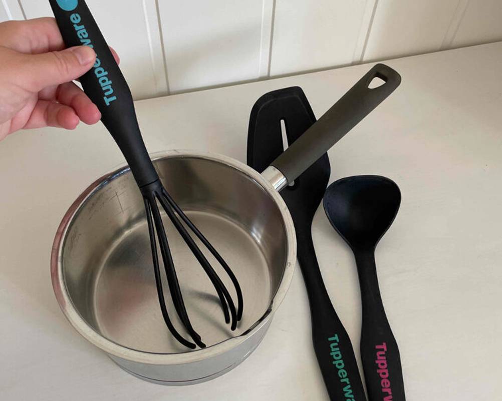 Küchenhelfer-Set-Test: Hand nutzt einen schwarzen Kunststoff-Schneebesen in einem Stieltopf, daneben liegen eine schwarze Kelle sowie ein Pfannenheber von Tupperware.