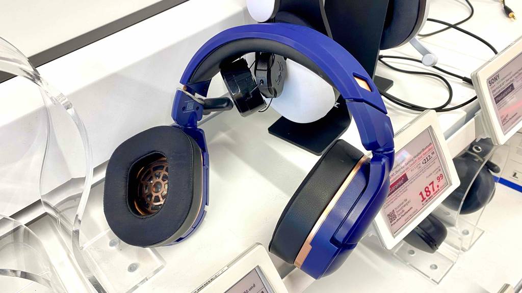 Im Test: Lilafarbiges PS4-Bluetooth-Headset lehnt an einem weißen Over-Ear-Kopfhörer auf einem Ausstellungsregal mit Preisschildern.