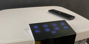 Ein weißer, hoch gebauter von oben betrachteter Büro-Luftreiniger im Test steht mit einem eingeschalteten Bedienpanel und einer schwarzen Fernbedienung auf einem Holzboden.