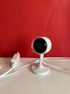 Auf einer hellen Fläche vor einer roten Wand steht eine Überwachungskamera mit Kabel und Ladegerät.