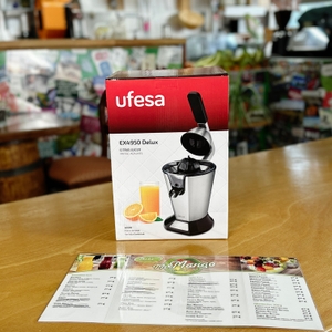 Die Verpackung der Ufesa Zitruspresse EX4950 steht auf einer Holztheke. Im Hintergrund ist ein Café mit einer bunt dekorierten Wand sichtbar.