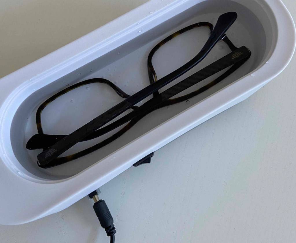 Weißer Ultraschall-Brillenreiniger im Test befindet sich ausgeschaltet inklusive Wasser und Brille mit schwarzem Gestell auf einer hellen Fläche.