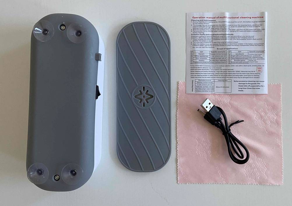 Ansicht der Aufstellfläche inklusive vier transparenter Saugnäpfe eines getesteten Ultraschall-Brillenreinigers neben seinem grauen Deckel, einem USB-Kabel, einem rosa Brillentuch und der Bedienungsanleitung.