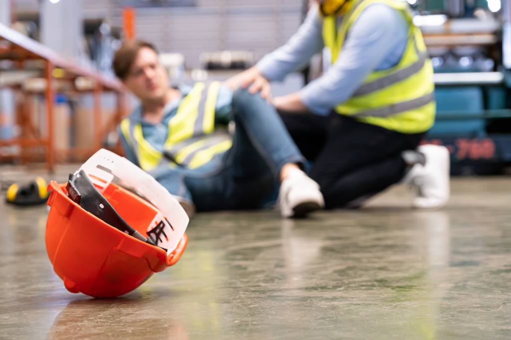 Eine verletzte Person sitzt am Boden einer Produktionshalle während sich ein anderer Arbeiter um sie kümmert.