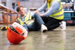 Unfallversicherungen im Test: Eine verletzte Person sitzt am Boden einer Produktionshalle während sich ein anderer Arbeiter um sie kümmert.