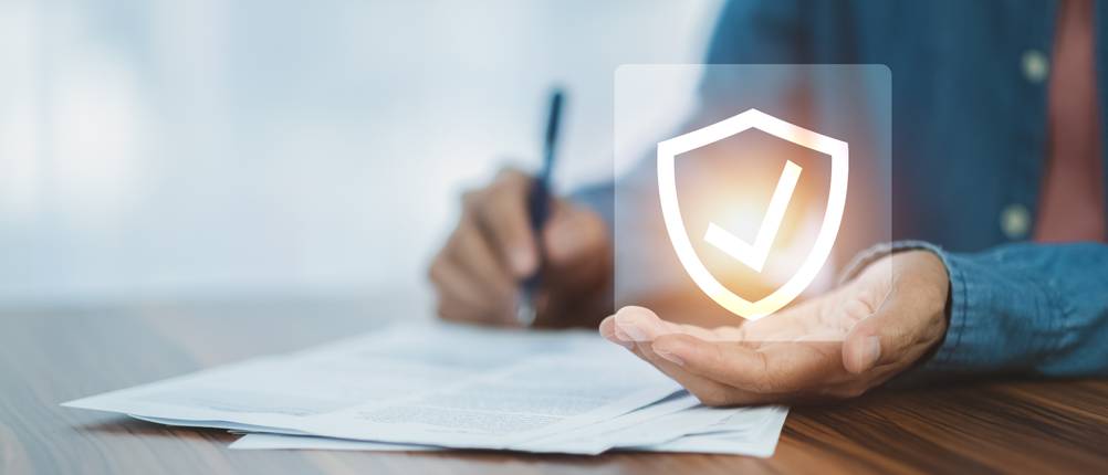 Unfallversicherung-Test: Eine Person unterschreibt ein Dokument, in seiner Hand ist ein Symbol zu sehen, welches für Sicherheit steht.