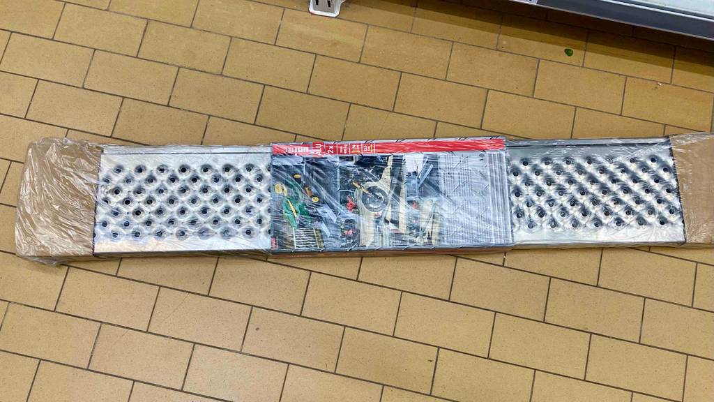 Eine in Kunststoff eingeschweißte Alu-Auffahrrampe liegt auf einem ockerfarbenen Fliesenbelag.