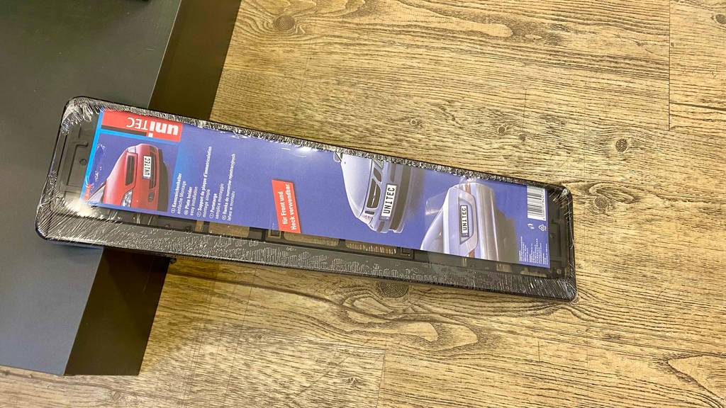 Im Test: Ein Kennzeichenhalter in Originalverpackung liegt quer auf einem Holzboden.