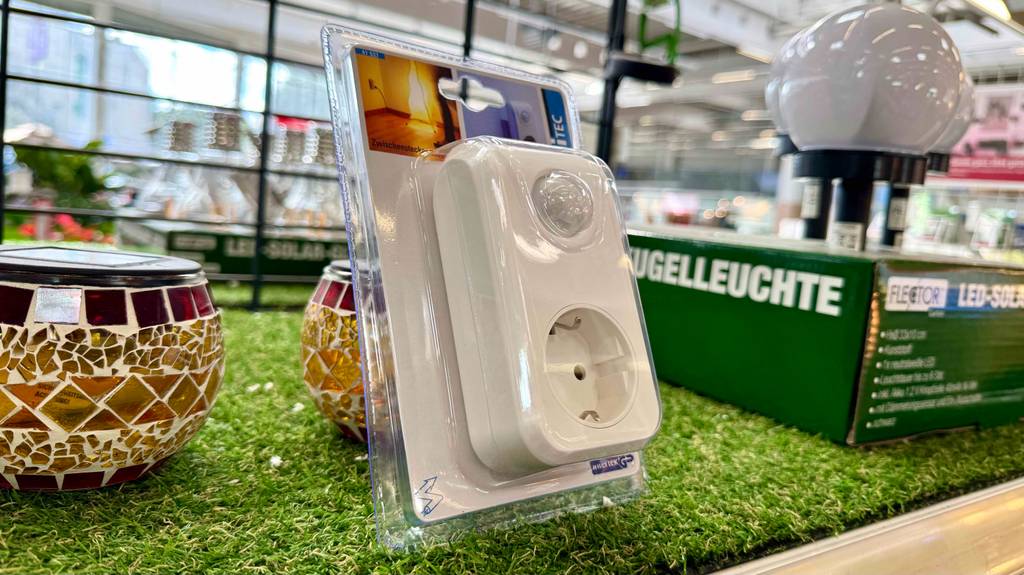 Das Nachtlicht mit Bewegungsmelder und Steckdose im Test befindet sich eingeschweißt in der transparenten Kunststoffverpackung auf einem Kunstrasen neben Solarleuchten im Baumarkt.