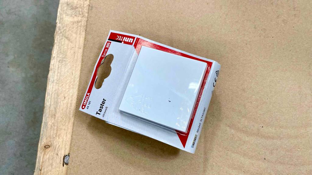 Im Test: ein Taster in ultraweißer, quadratischer Ausführung in seiner Verpackung auf einer hellbraunen Oberfläche.