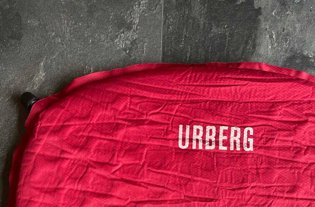 Getestet: Eine selbstaufblasbare Isomatte in Rot inklusive schwarzem Ventil und der weißen Aufschrift „URBERG“ liegt auf einer grauen Oberfläche.