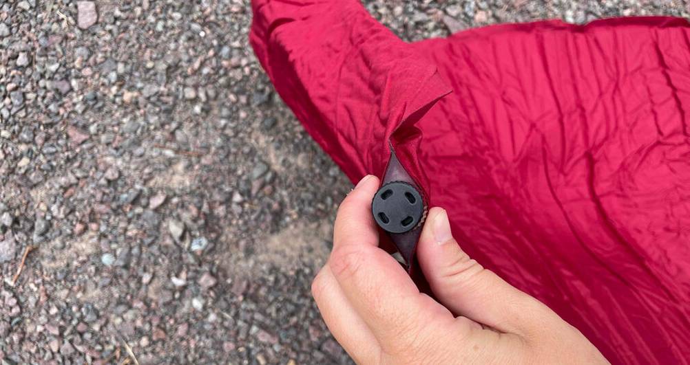 Selbstaufblasbare Isomatten im Test: Hand hält eine rote Isomatte, mit Fokus auf das schwarze Ventil, im Hintergrund ein Schotterboden.