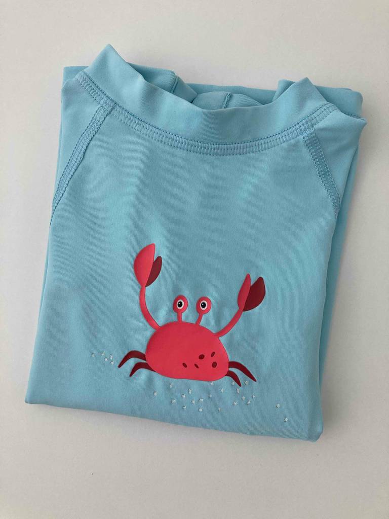 Das getestete Kinder-UV-Shirt in Hellblau mit einem mittig bedruckten roten Krebs liegt zusammengefaltet auf einer weißen Oberfläche.
