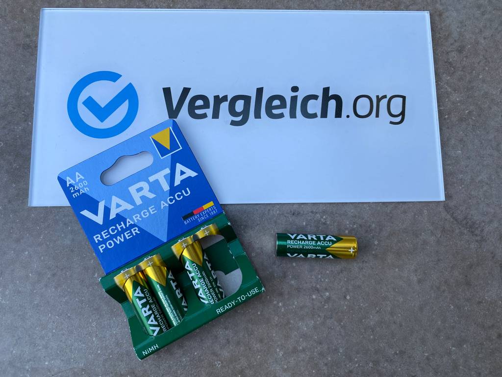 Vergleich.Org Logo und Varta AA-Akkus liegen auf dem Boden. Ein Akku wurde aus der Verpackung genommen und daneben gelegt.