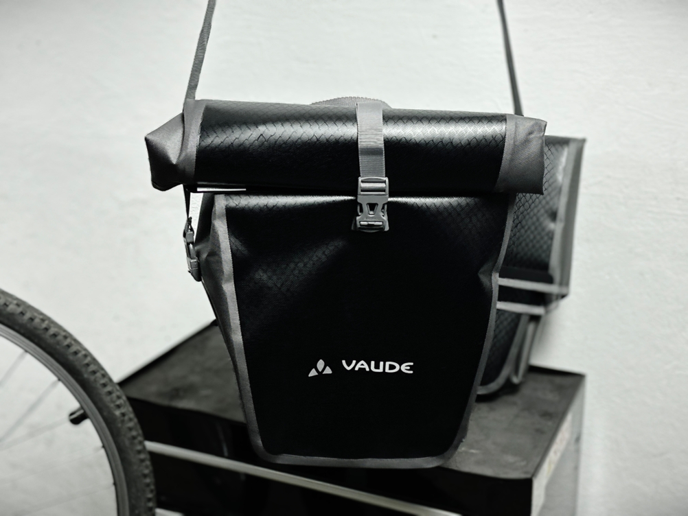 Die Vaude-Aqua-Back-Tasche hängt an einem Schultergurt und wird als Tragetasche verwendet. Im Hintergrund ist ein Fahrradreifen zu sehen.