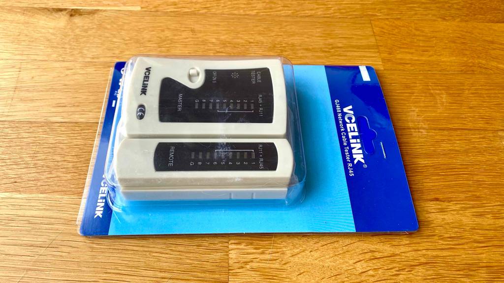 Im Test: Ein Kabeltester RJ45 der Marke VCLiNK liegt in seiner blauen Verpackung hinter durchsichtigem Kunststoff auf einem Holzuntergrund.