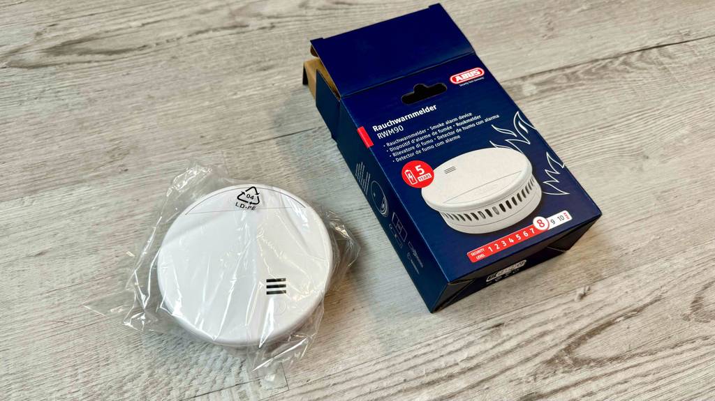 Ein getesteter Abus-Rauchmelder liegt neben der Originalverpackung auf einer Holzfläche.