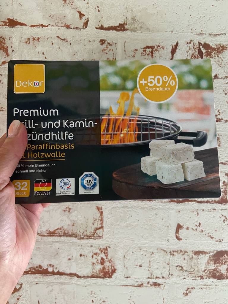 Eine Hand hält die getesteten Anzünder in der DEKOR-Packung mit Anzünder-Test: Eine Packung Grill- und Kaminzündhilfen vor einer weißen Ziegelmauer.
