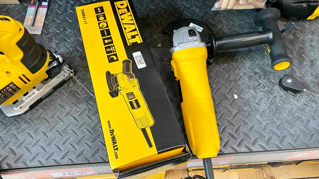 Ansicht von oben auf einen gelben DeWalt Winkelschleifer im Test, der neben seiner Verpackung auf einem grauen Metallregal im Verkaufsbereich neben anderen Geräten ausgestellt ist.