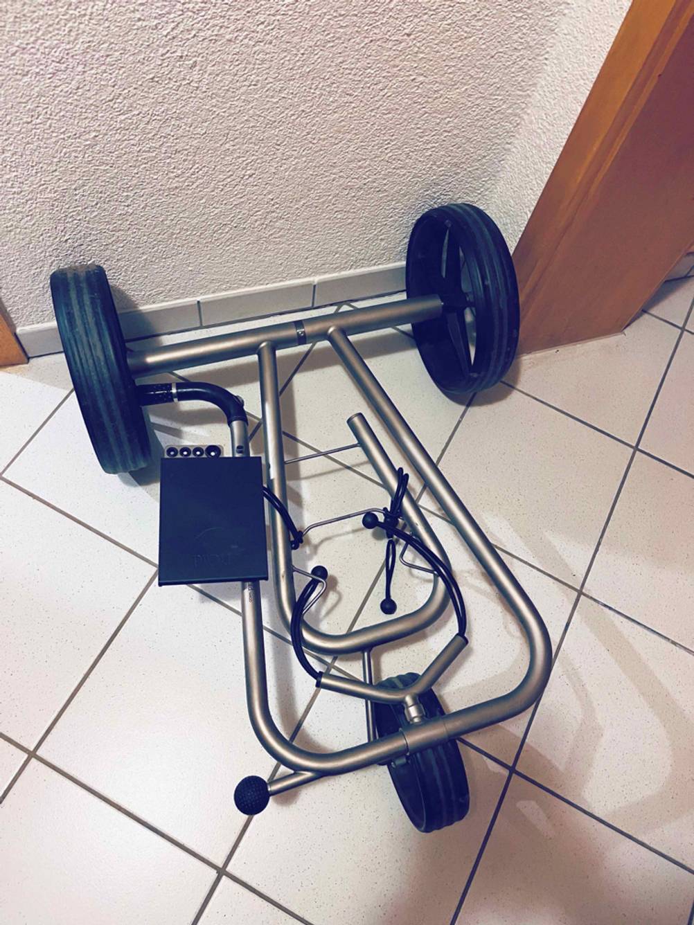 Gesamtansicht von oben eines auf weißen Fliesen liegenden Aluminium-Gestells inklusive schwarzer Räder eines getesteten Golf-Trolleys.