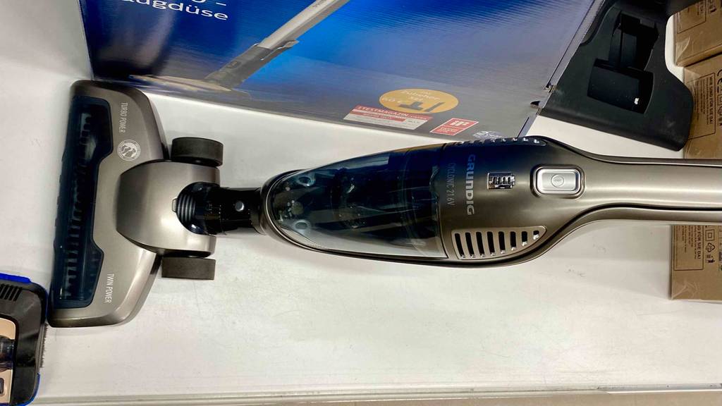 Grundig-Akku-Staubsauger im Test: Ansicht von oben auf einen silberfarbenen Stielstaubsauger, mit Fokus auf den Bürstenaufsatz und seinen transparenten Staubfangbehälter neben einer blauen Verpackung auf einem weißen Verkaufsregal.