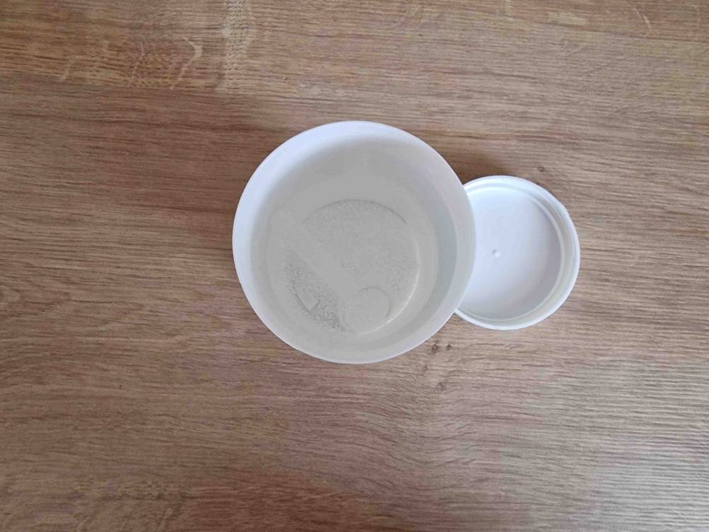 Im Test: Magnesiumcitrat-Pulver in einer geöffneten weißen Kunststoff-Dose inklusive Dosierlöffel und Schraubdeckel von oben, auf Holzfläche.
