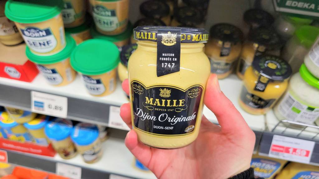 Maille-Senf getestet: Eine Hand hält ein Glas mit originalem Dijon-Senf vor einem Supermarktregal.
