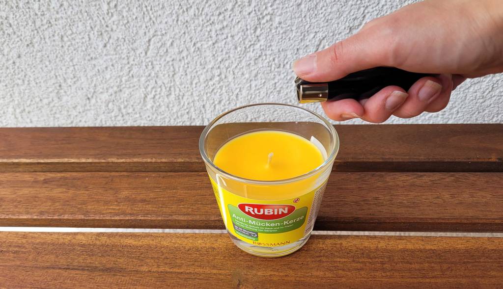Anti-Mücken-Kerzen-Test: Hand hält ein Feuerzeug über eine gelbe Kerze im Glas, das auf einer Holzfläche steht.