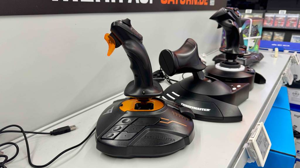 Der getestete Thrustmaster-Joystick in Schwarz mit orangefarbenen Highlights steht vor einer Reihe weiterer Joysticks auf einem weißen Ausstellungsregal.