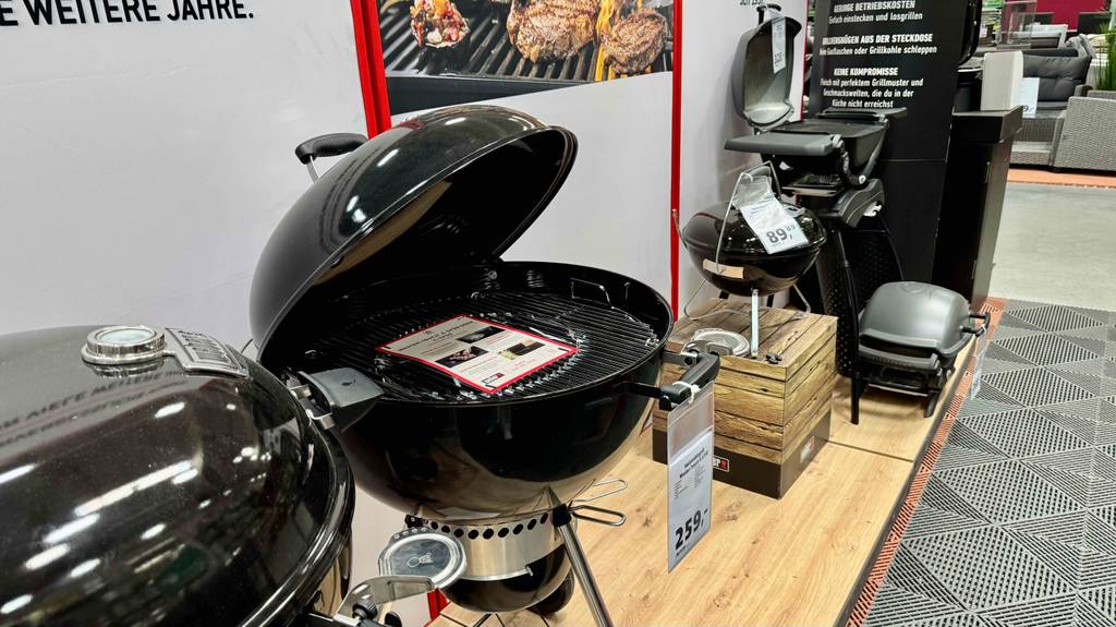 In einem Verkaufsbereich mit verschiedenen Holzkohlegrills befindet sich der schwarze, runde, getestete Weber-Holzkohlegrill inklusive Standfüße.