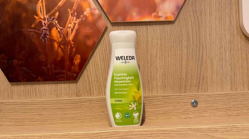 Eine Kunststoffflasche mit WELEDA Bodylotion im Test steht auf einem Regal in Holzoptik.