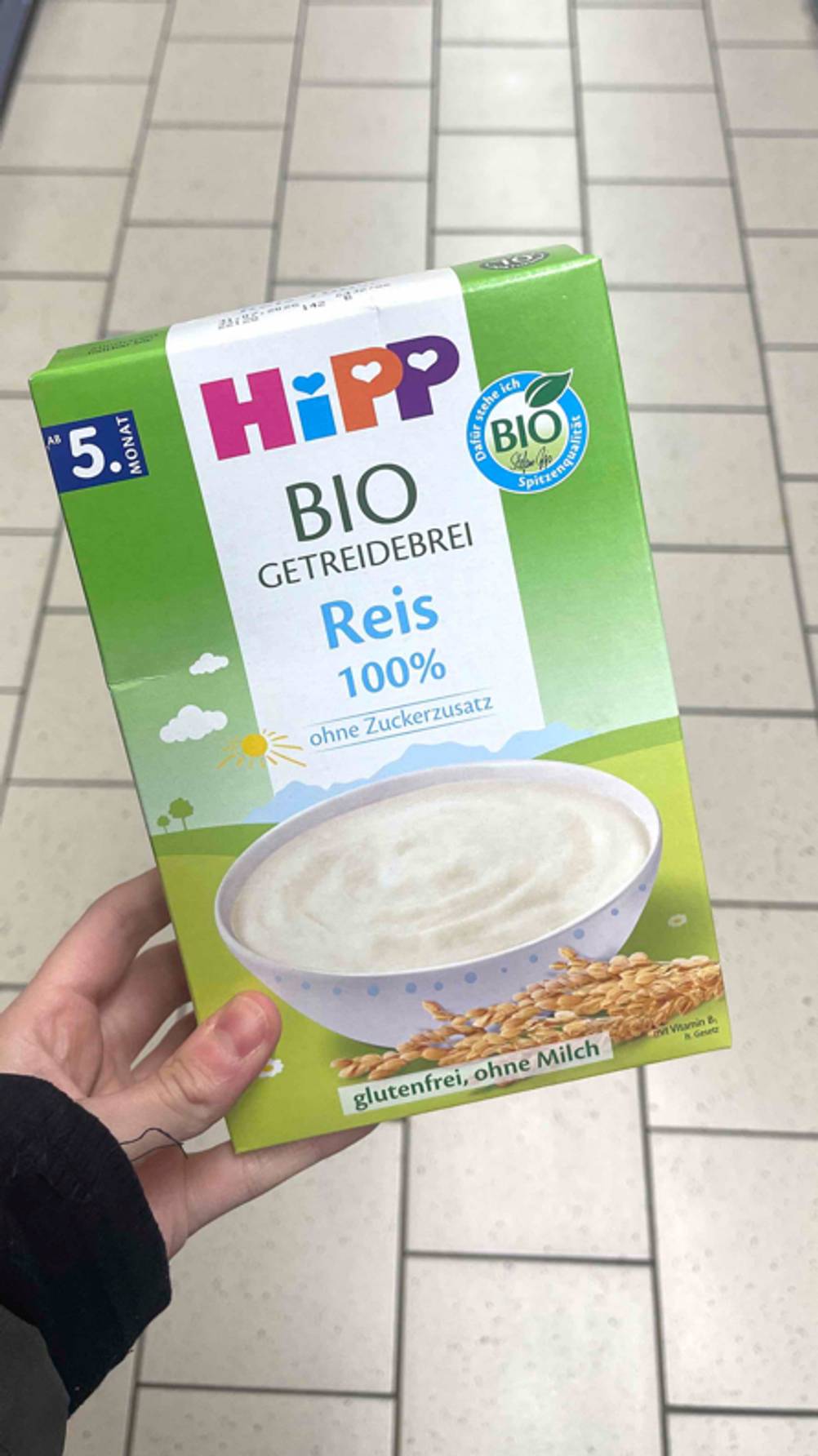 Getreidebrei im Test: Eine Hand hält die grüne 200-g-Packung Reisbrei von Hipp über einem weißen Fliesenboden.