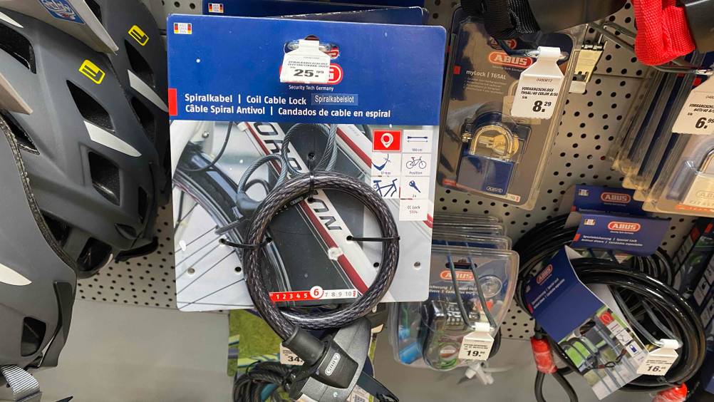 ABUS-Fahrradschloss-Test: ein schwarzes Spiralkabel-Schloss hängt mit seiner blauen Verkaufspappe an einem Verkaufsregal zusammen mit weiterem Fahrradzubehör.