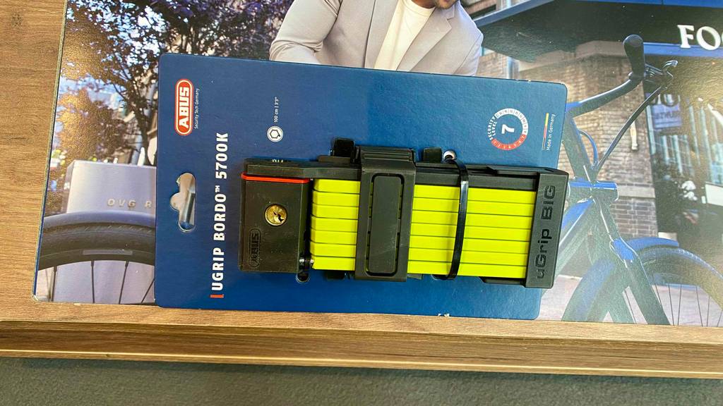 Ein gelbes Abus-Faltschloss im Test liegt in seiner blauen Verpackung vor einem Plakat mit einem Fahrradfoto.