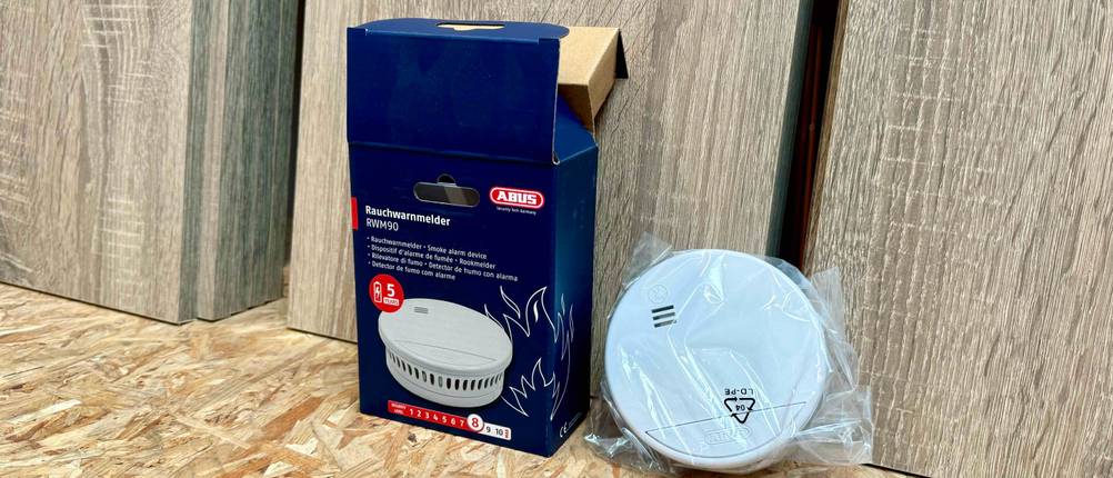 Ein Abus-Rauchmelder im Test lehnt neben seiner Originalverpackung an einer Holzfläche.