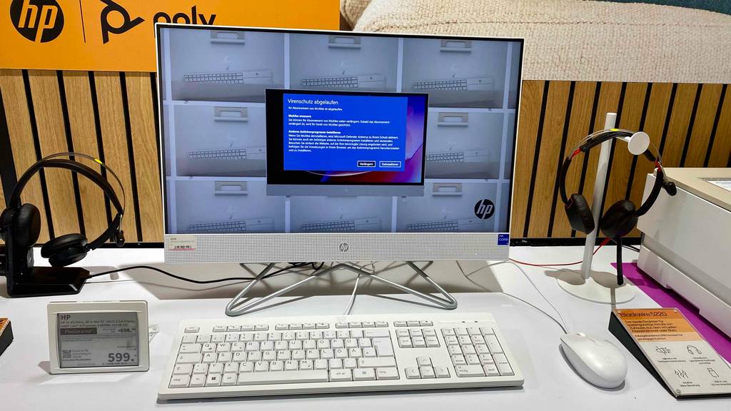 Weißer HP All-in-One-PC im Test befindet sich zwischen ausgestellten schwarzen Kopfhörern und neben einem weißen Drucker auf einem weißen Verkaufstisch inklusive Preisschild.
