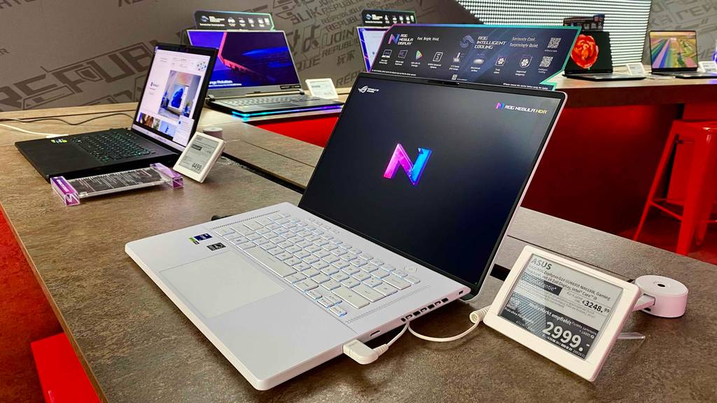 Man sieht die seitliche Ansicht über Eck auf einen silberfarbenen Asus-Gaming-Laptop auf einem braunen Ausstellungstisch neben anderen Laptops.