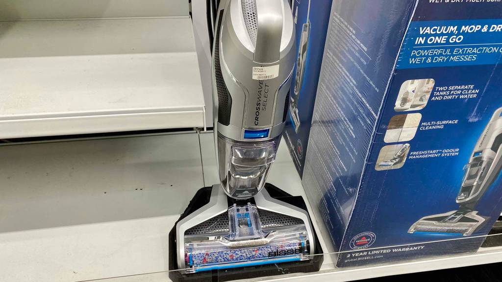 Vorderansicht von oben auf einen metallfarbenen, beutellosen Bissell-Staubsauger im Test, der mit einer transparenten Bodendüse ausgestattet ist und neben seiner blauen Originalverpackung steht.