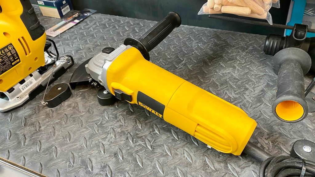 DeWalt-Winkelschleifer-Test: ein gelbes Modell liegt mit Fokus auf den rutschfesten, schwarzen Griff auf einem grauen Metallregal im Verkaufsbereich zwischen weiteren Geräten.
