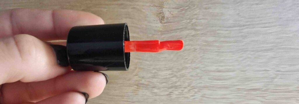 Gel-Nagellack-Test: Hand hält einen Nagellack-Pinsel mit rotem Gel vor einem Holzhintergrund.