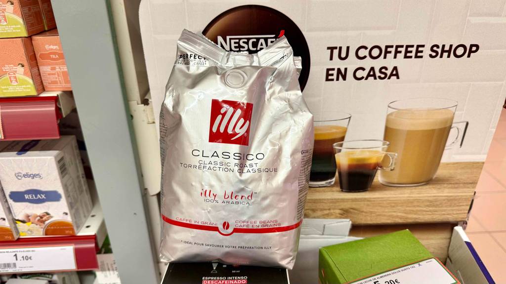 Eine silberfarbige Tüte mit den getesteten illy-Kaffeebohnen steht vor einem Plakat mit gefüllten Kaffeetassen.